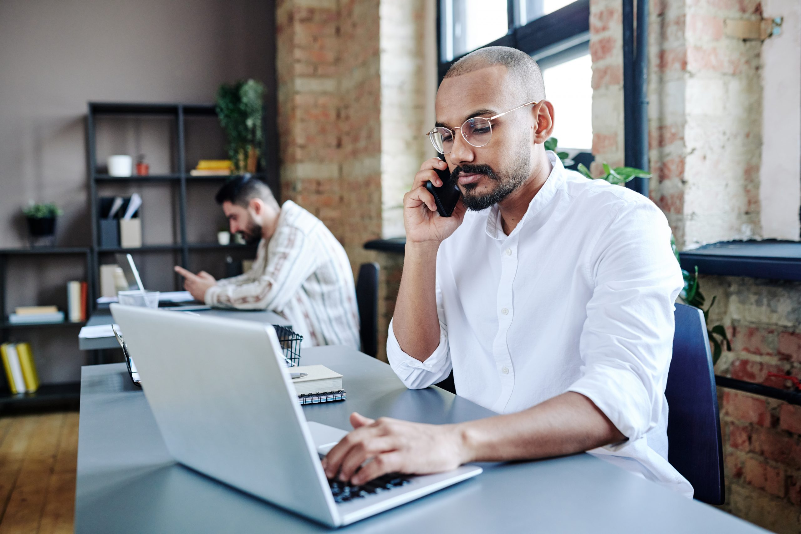 Busy young employee in casualawear talking on mobile phone by workplace